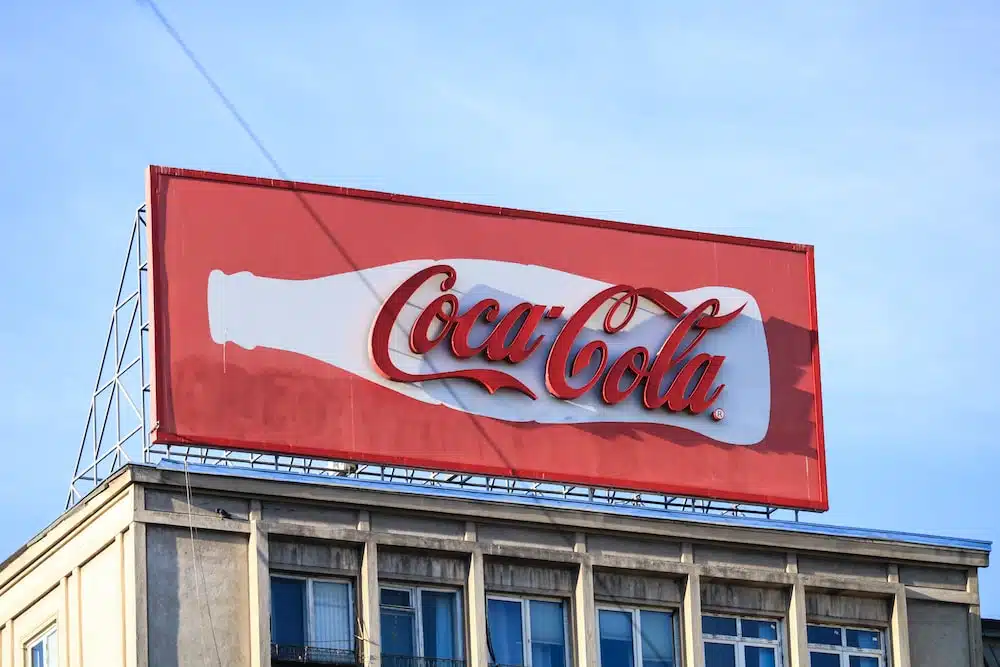 A large red Coca-Cola billboard featuring the iconic white contour bottle and script logo mounted on a city building. This classic outdoor advertisement stands against a clear blue sky, showcasing global brand recognition.