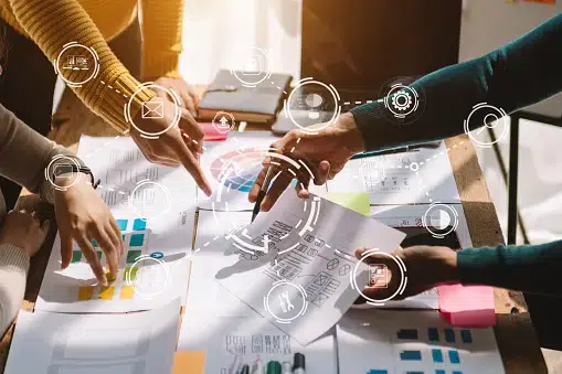 A diverse team of professionals collaborating around a table covered in design sketches and color palettes. Digital icons representing email, settings, and communication are overlaid to signify integrated marketing workflows.