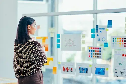 A marketing professional stands before a glass wall covered in color swatches, charts, and sticky notes while planning a project. She appears deep in thought, highlighting the analytical and creative process of brand development.