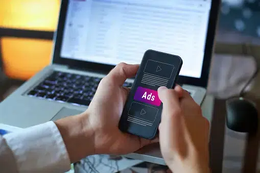 A close-up shot of a person holding a smartphone displaying a purple "Ads" notification over a video interface. A laptop is visible in the background, illustrating the ubiquity of cross-platform digital advertising.