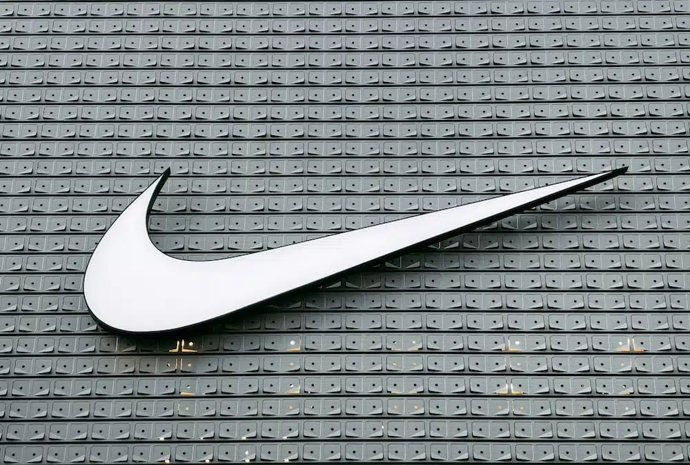 A large white Nike Swoosh logo mounted on a modern grey textured wall with a repetitive geometric pattern. The minimalist design emphasizes the brand's athletic identity and contemporary architectural style.