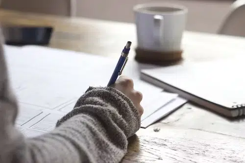 A close-up shot of a person's hand using a blue pen to draw layout sketches or wireframes on a sheet of paper. This captures the early stages of creative design, planning, or architectural drafting at a sunlit table.