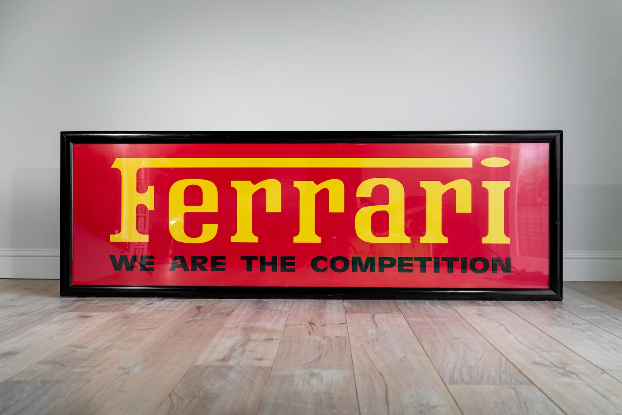 Framed Ferrari sign on a wooden floor with bold red and yellow branding and the slogan 'We Are the Competition,' reflecting a famous slogan tied to luxury and performance.