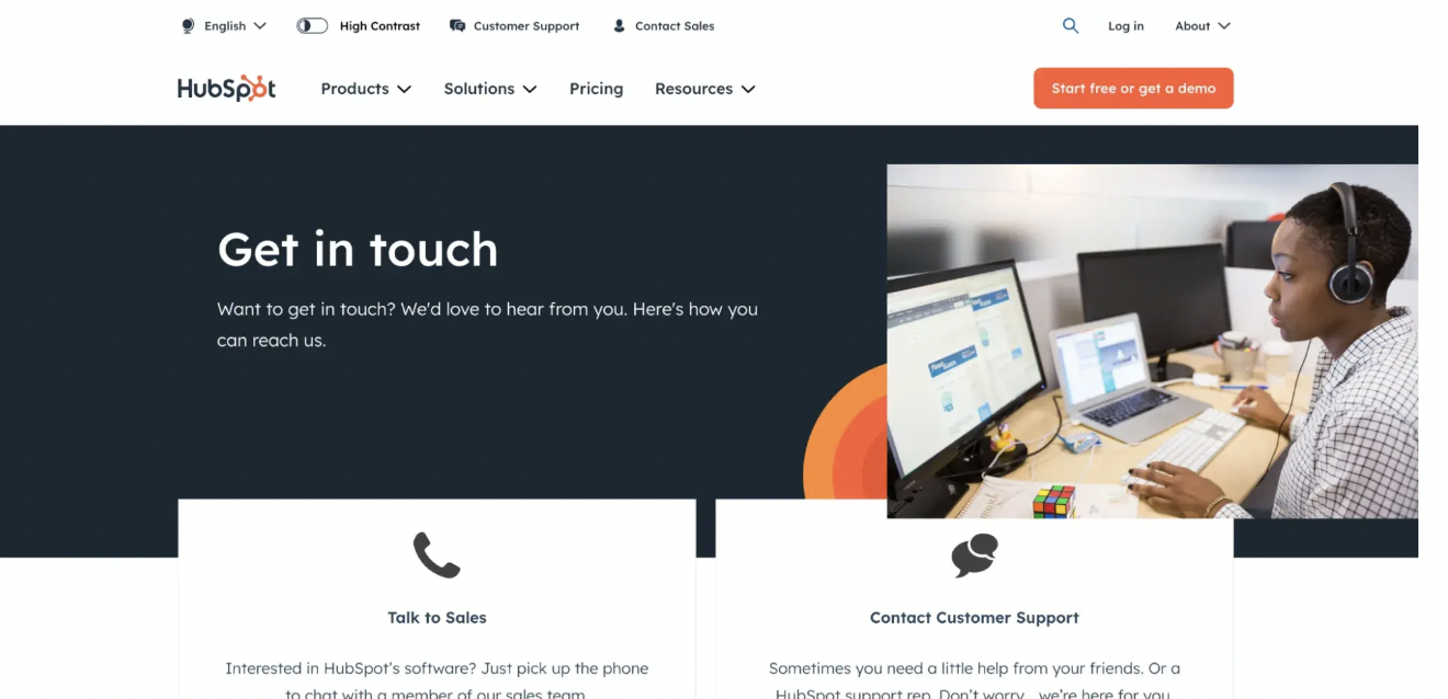 HubSpot’s contact page features a dark background with the heading “Get in touch,” offering users the option to speak with sales or contact customer support. On the right, a photo shows a woman wearing a headset working on multiple monitors in a modern office. Below the image are two clearly labeled contact options for sales inquiries and customer support help.