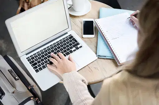 Woman working at a round table with a laptop, notebook, smartphone, and suitcase, writing notes while typing.