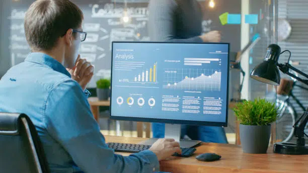 Man sitting at a desk analyzing charts and graphs displayed on a large computer monitor in a modern office setting.