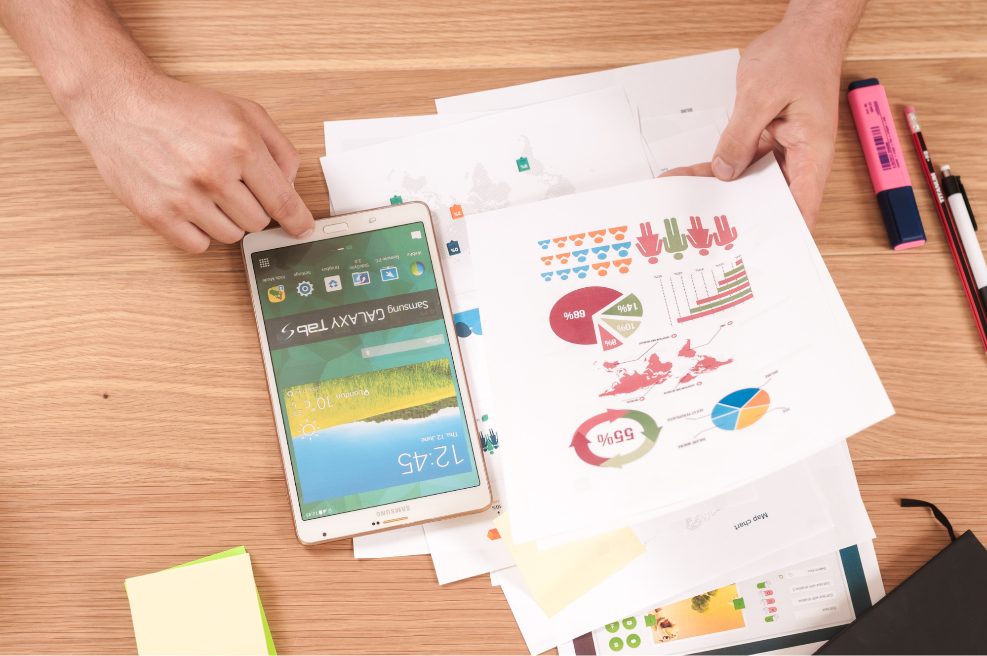 Person reviewing colorful printed charts and graphs on a wooden desk with a Samsung Galaxy Tab, pens, highlighter, and sticky notes.