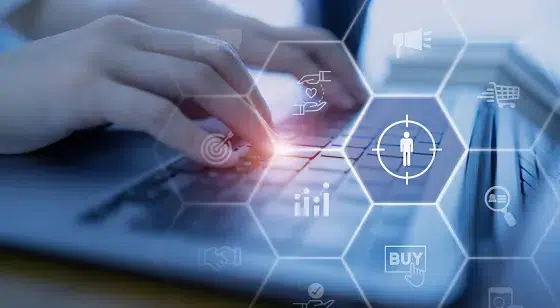 Hands typing on a laptop keyboard with digital hexagon icons overlayed, including a target audience symbol, shopping cart, and buy button, representing digital marketing and audience targeting.