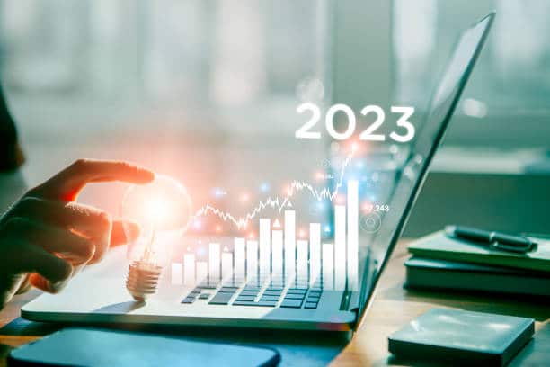 Hand holding a glowing lightbulb near a laptop with digital bar charts, graphs, and the year "2023" representing innovation and future trends in AI marketing.