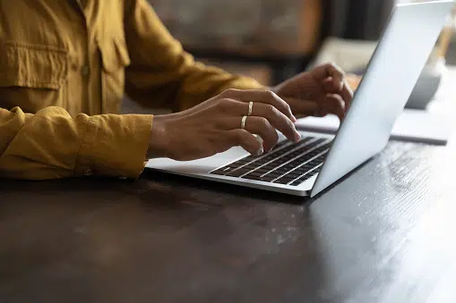 Person wearing a mustard yellow shirt typing on a laptop at a dark wooden desk.