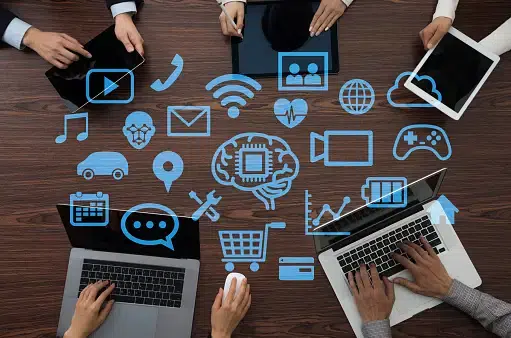 People working on laptops and tablets at a table with digital icons overlaid, representing technology, communication, e-commerce, and data analysis tools.