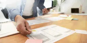 Person working on UX design with smartphone in hand, placing paper wireframe templates on a desk with sticky notes.