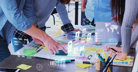 Team collaborating around a table with sticky notes, charts, and a laptop, using holographic digital interface elements to plan a UX strategy.