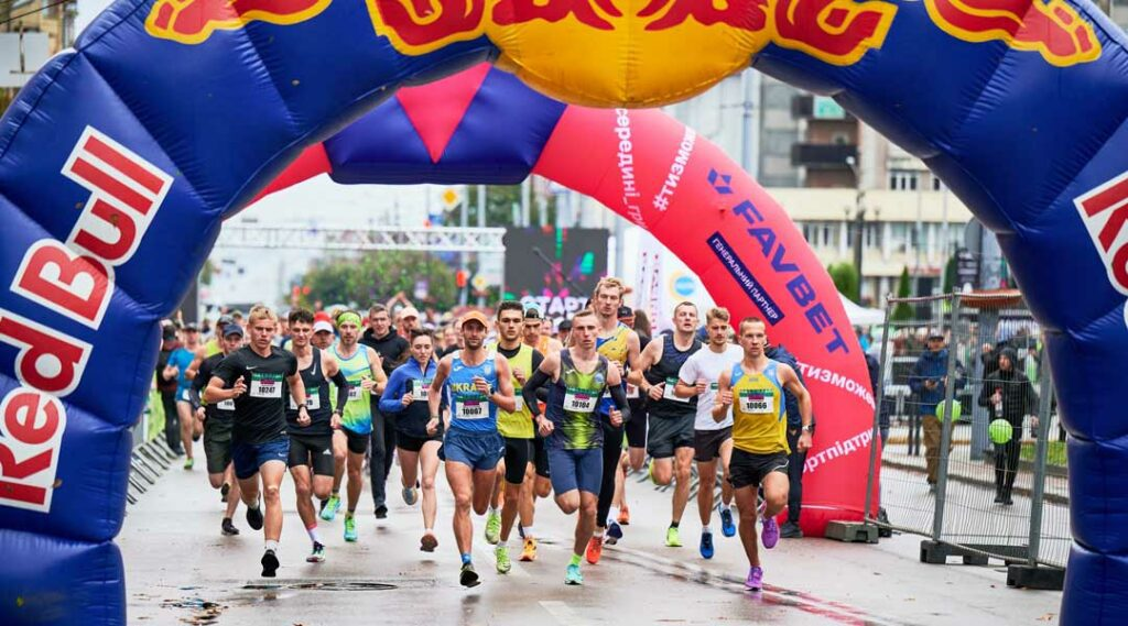A group of marathon runners takes off from the starting line beneath a large Red Bull inflatable arch during a city race.