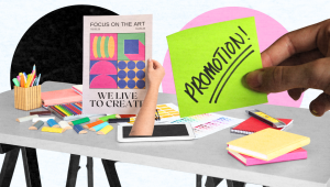 A hand holds a green sticky note that says PROMOTION! over a desk scattered with art supplies, notebooks, colored pencils, and a poster reading FOCUS ON THE ART. WE LIVE TO CREATE—perfect for creative brand promotion.
